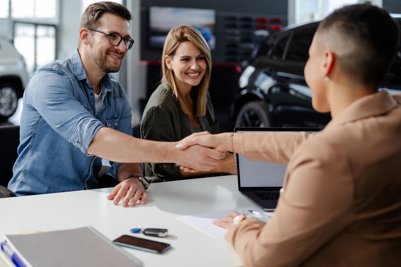 A couple shaking hands with the car salesperson sitting at his desk.