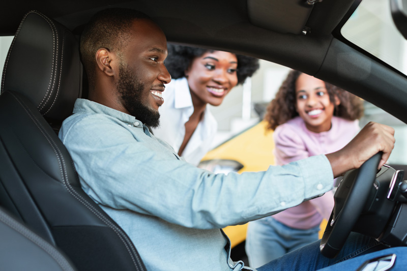 A family smiling standing outside of a car while the father sits inside in the driver's seat.