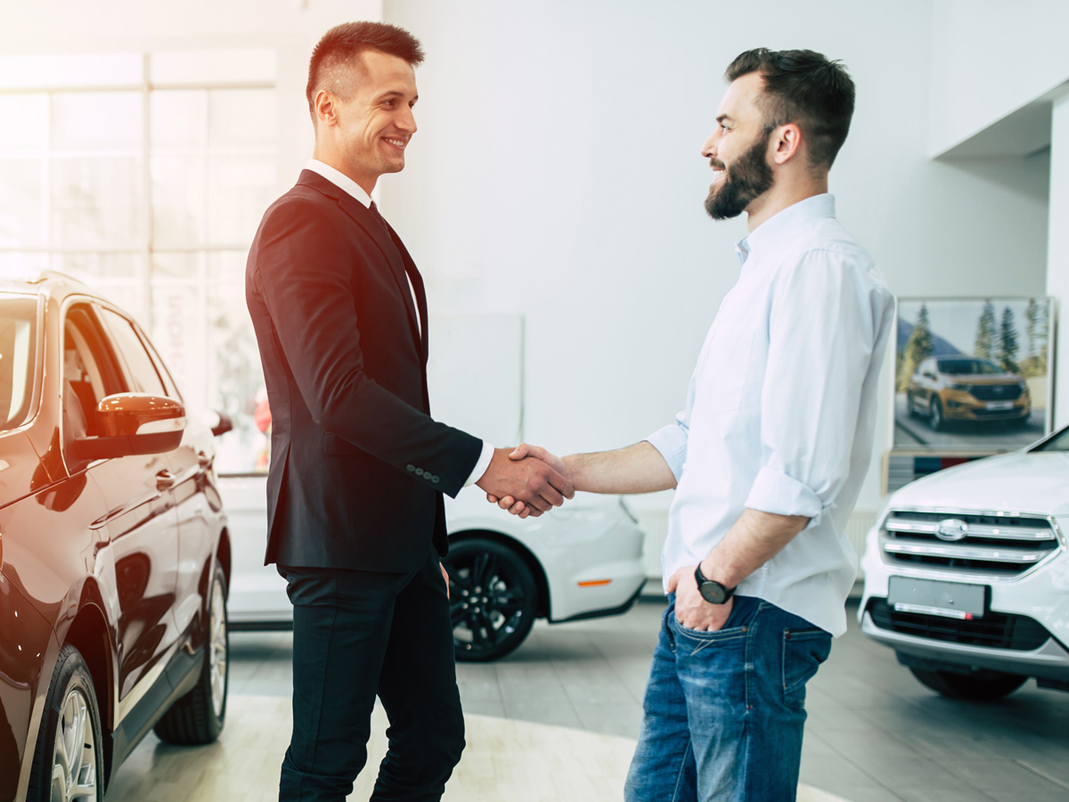 A car salesperson at the dealership shaking hands with a smiling customer.