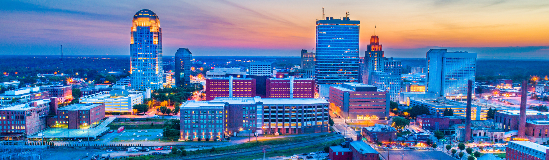 The downtown skyline of Winston-Salem, North Carolina, featuring iconic landmarks such as the Wells Fargo Center (left) and the Winston-Salem Tower (center-right) at dusk.