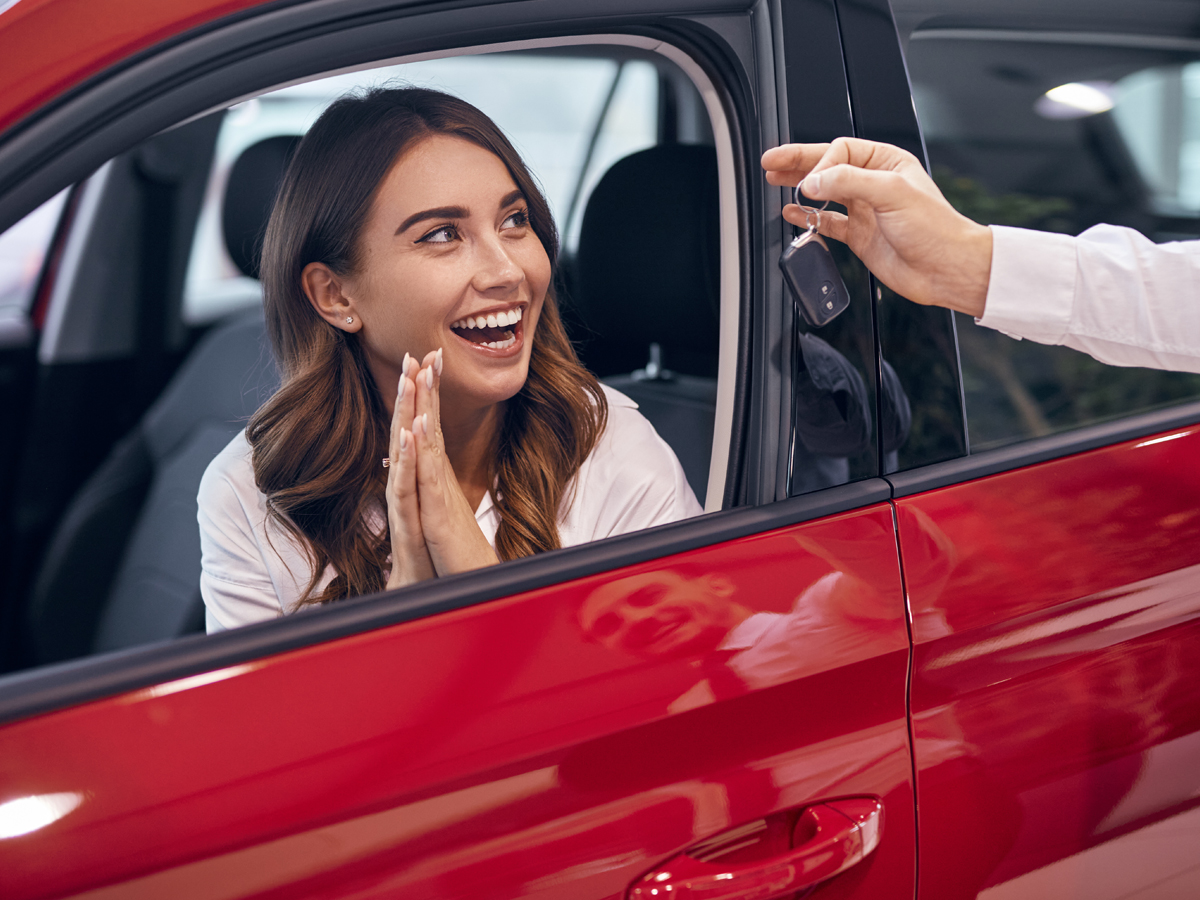 A smiling woman in a car getting car keys handed to her through the car's window.