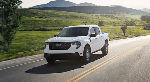 A white Ford Maverick pickup truck driving on a road by open fields.
