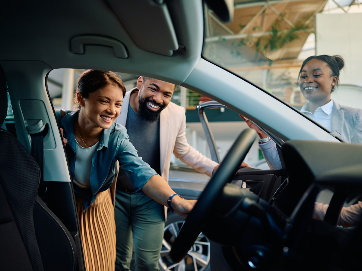 A car salesman showing the inside of a vehcile to a smiling couple leaning in to the vehicle's open door.