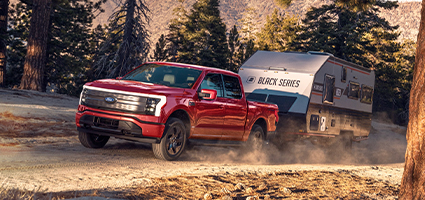 A red Ford F-150 Lightning hauling a trailer behind it on a dusty mountain road.