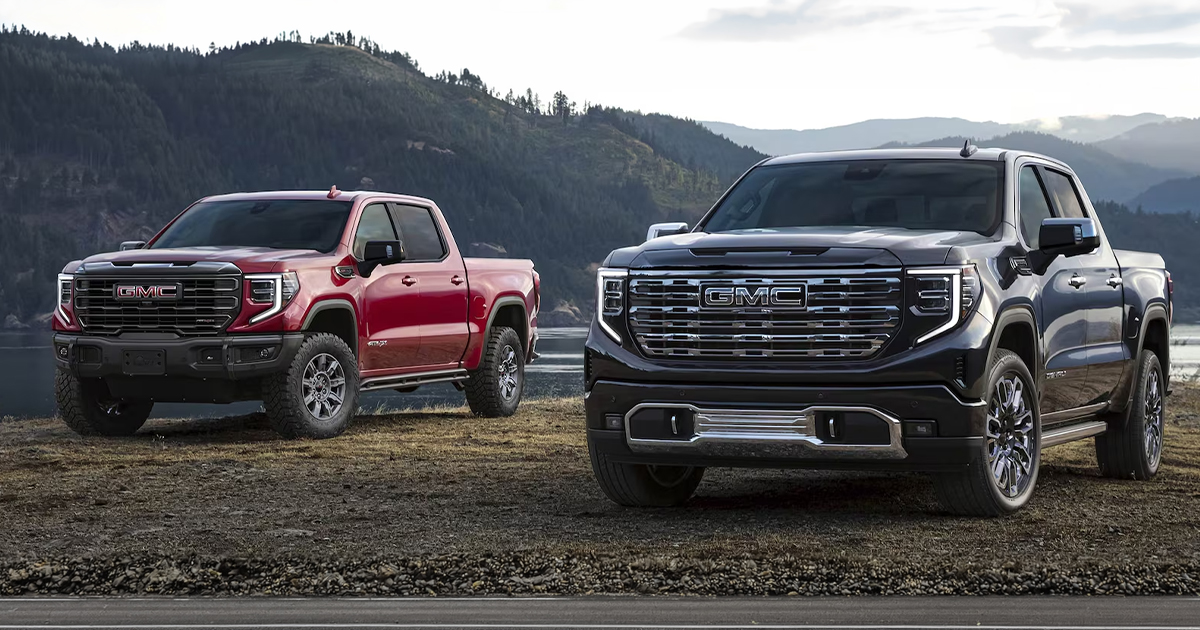 A GMC Sierra AT4X in red on the left and a luxury-focused GMC Sierra Denali Ultimate in dark trim on the right parked by a lake.