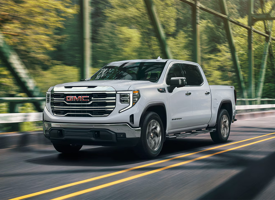 A GMC Sierra 1500 pickup truck, featuring a prominent chrome grille and a white exterior finish driving across a bridge.