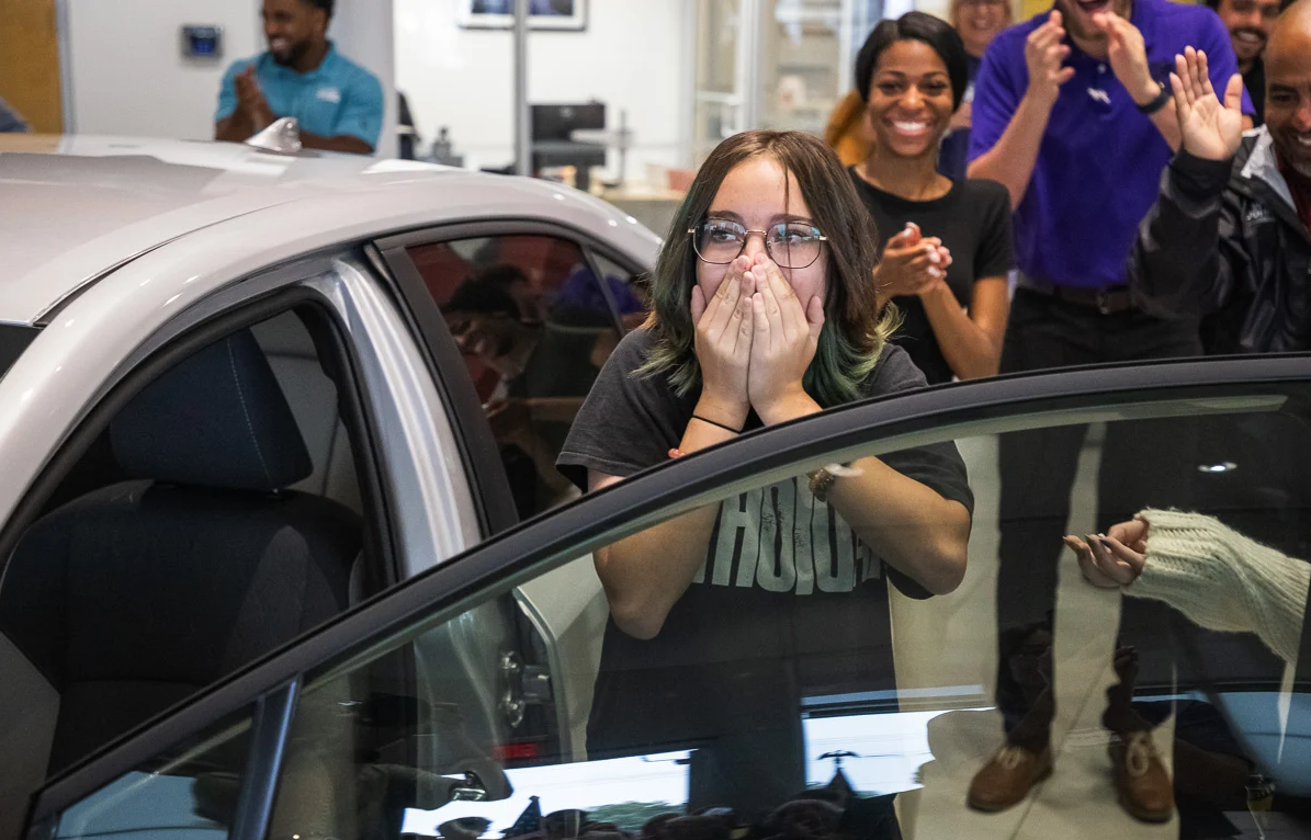 A shocked student standing beside a brand new car they won at the dealership while staff and friends cheer.